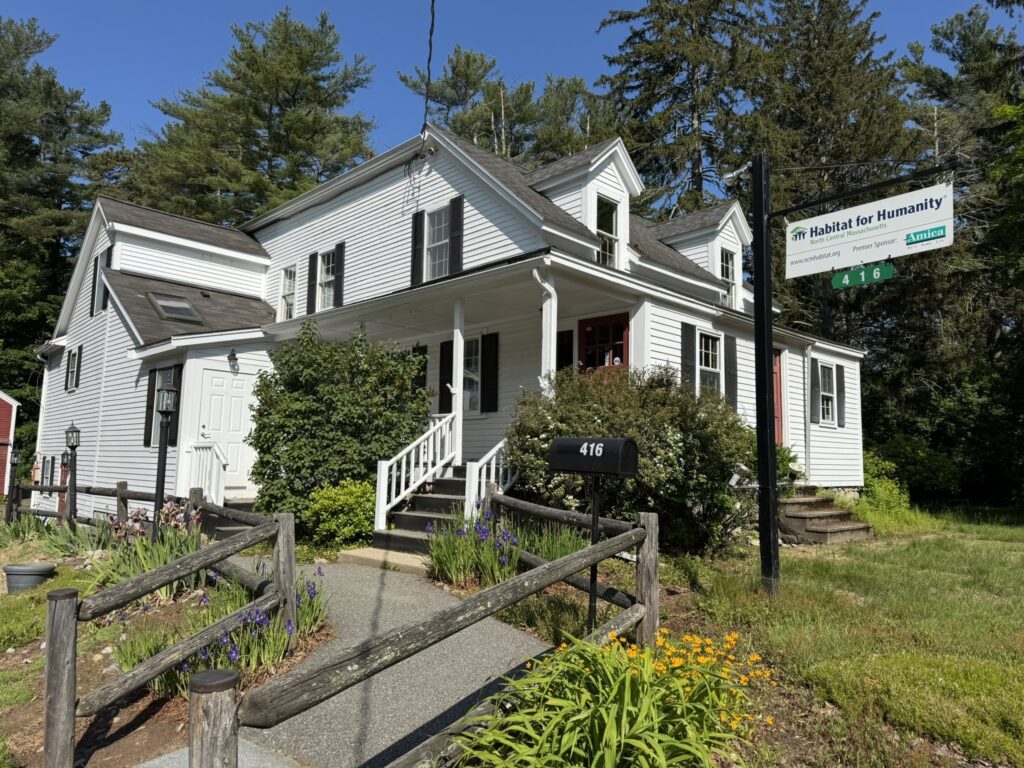 A white house with dormers and a porch. There is a sidewalk leading up to the front steps.