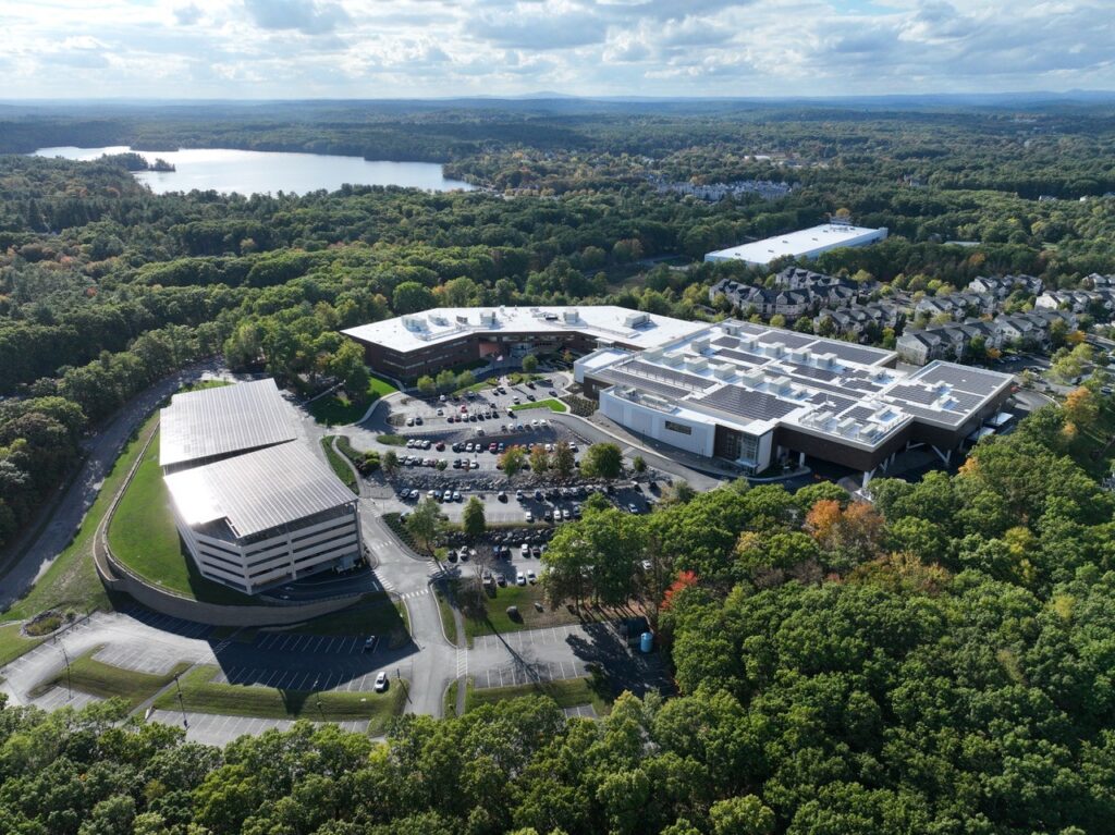 An aerial view of a very large low building. A pond sparkles in the background and trees are in the foreground.