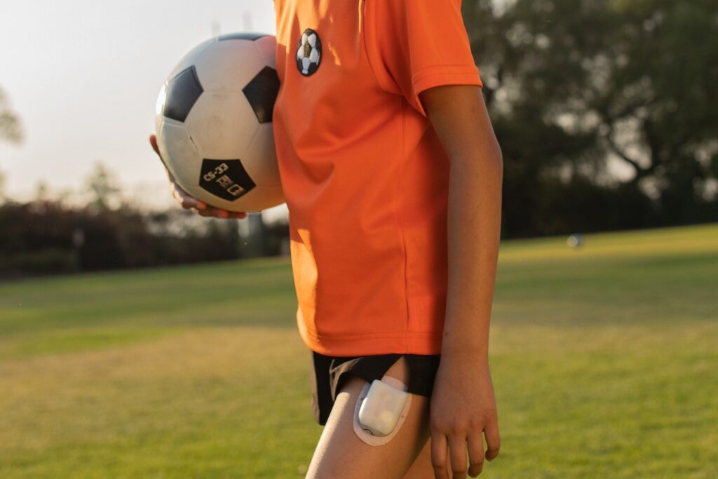 A picture of a body (no head) wearing a soccer jersey and holding a soccer ball. An OmniPod patch is on their thigh.