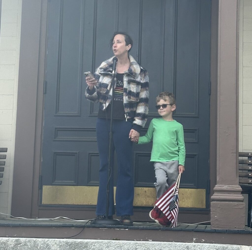 A woman speaks into the mic. She's holding the hand of a young boy who is carrying American flags.