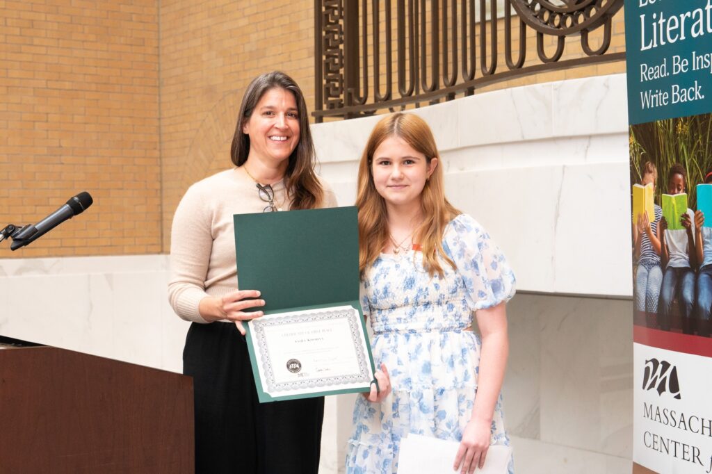 A blond girl in a blue flowered dress sands with a brown-haired woman. The woman holds a citation.