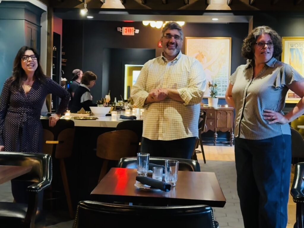 Two women and a bearded man stand in a restaurant. In the background two people are setting up.
