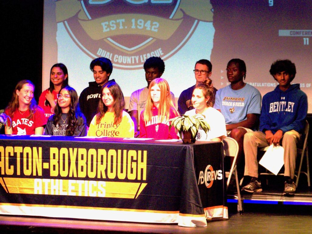 A group of students wearing college t-shirts and sweatshirts sit ata table. The tablecloth says Acton-Boxborough Athletes.