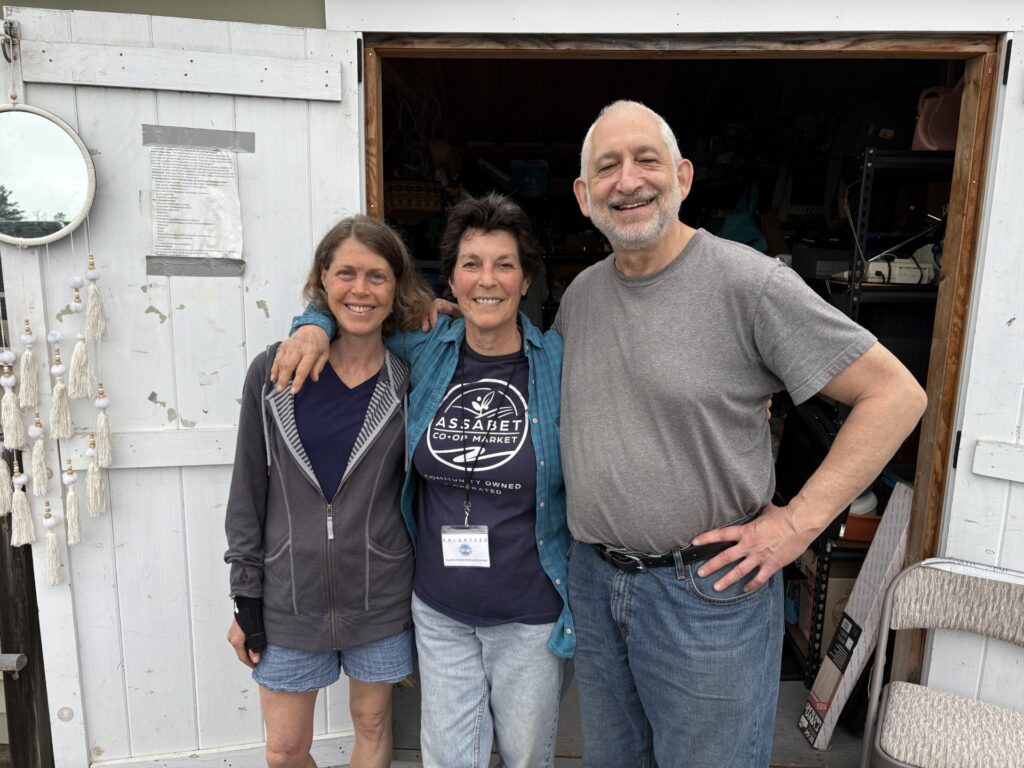 Two women and a bearded man stand with their arms on each others shoulders in front of an open white door.