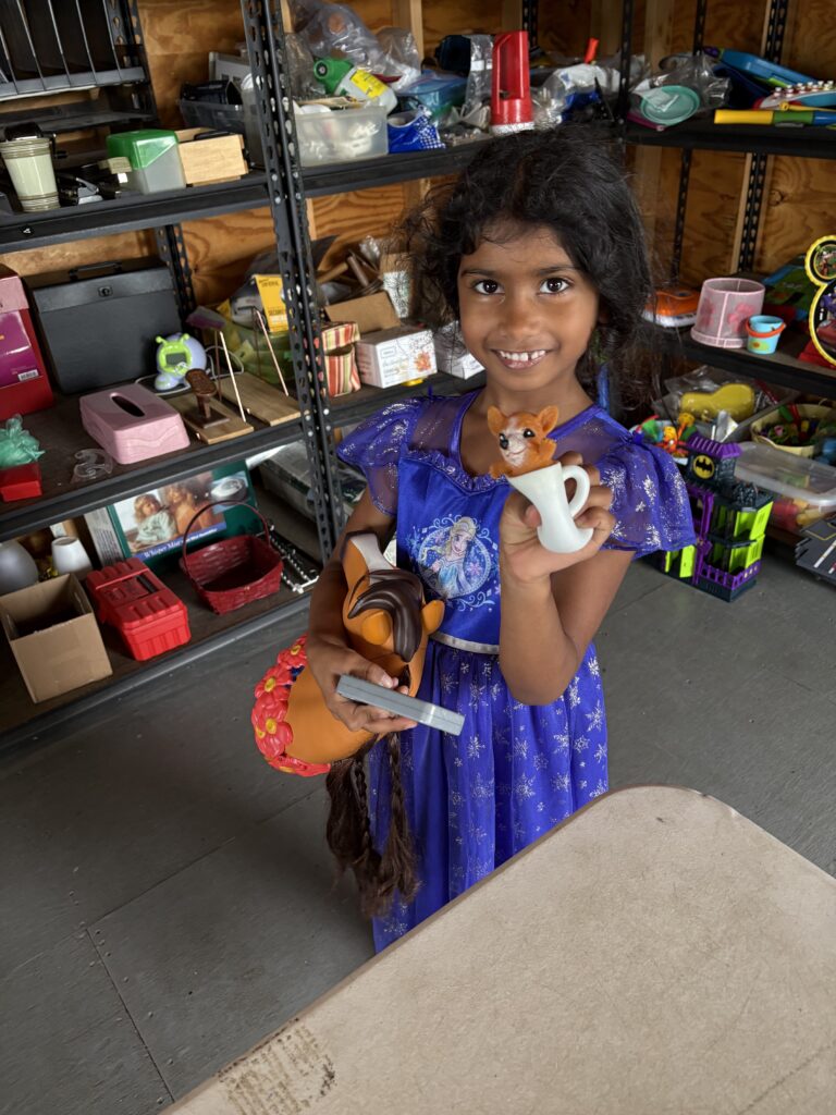 A young girl in a blue dress shows off her new squeezy toy. A toy horsehead is under her arm.