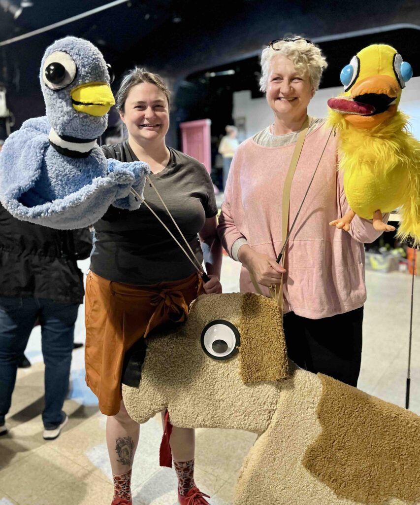 Two female actors hold soft, colorful puppets. On the left, Rosemary Ouellet holds The Pigeon; on the right, Wendy Blizard Eldredge holds Duckling and Puppy. They are rehearsing for a family-friendly musical at Theatre III in Acton.