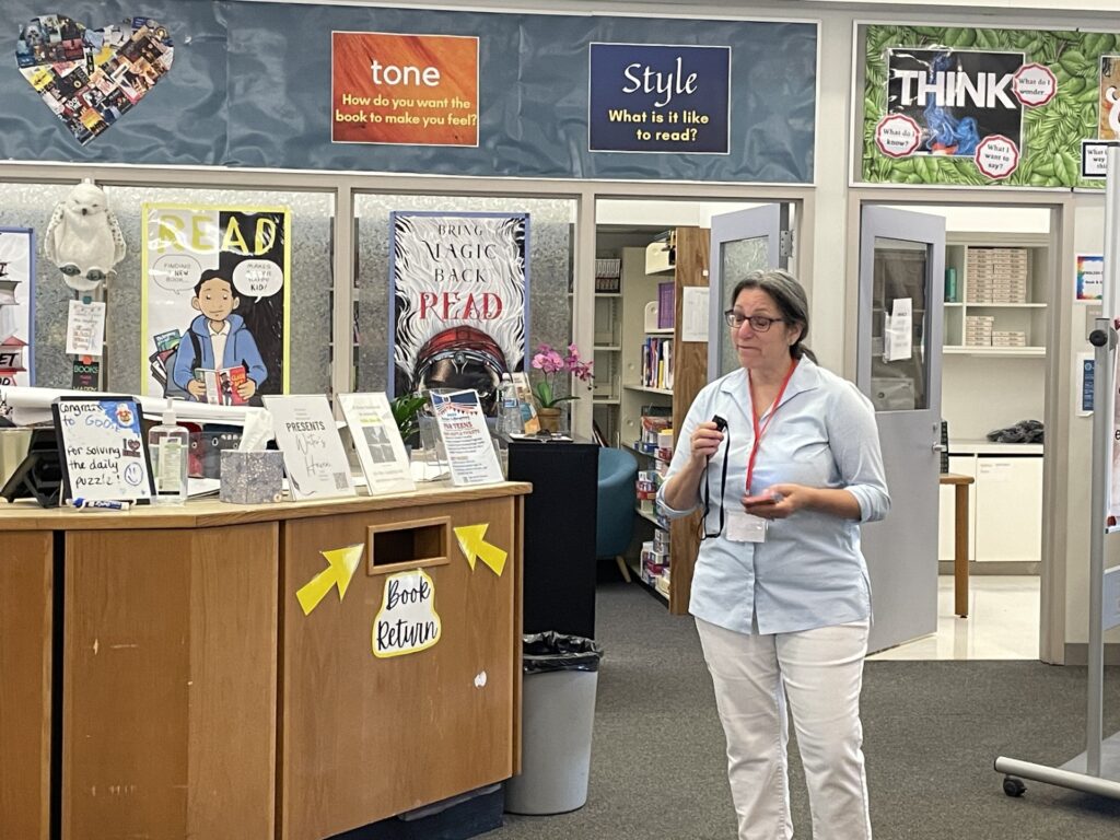 A woman in a blue shirt holds a small microphone. She is standing next to the library Book Return slot and there many posters up on the wall.