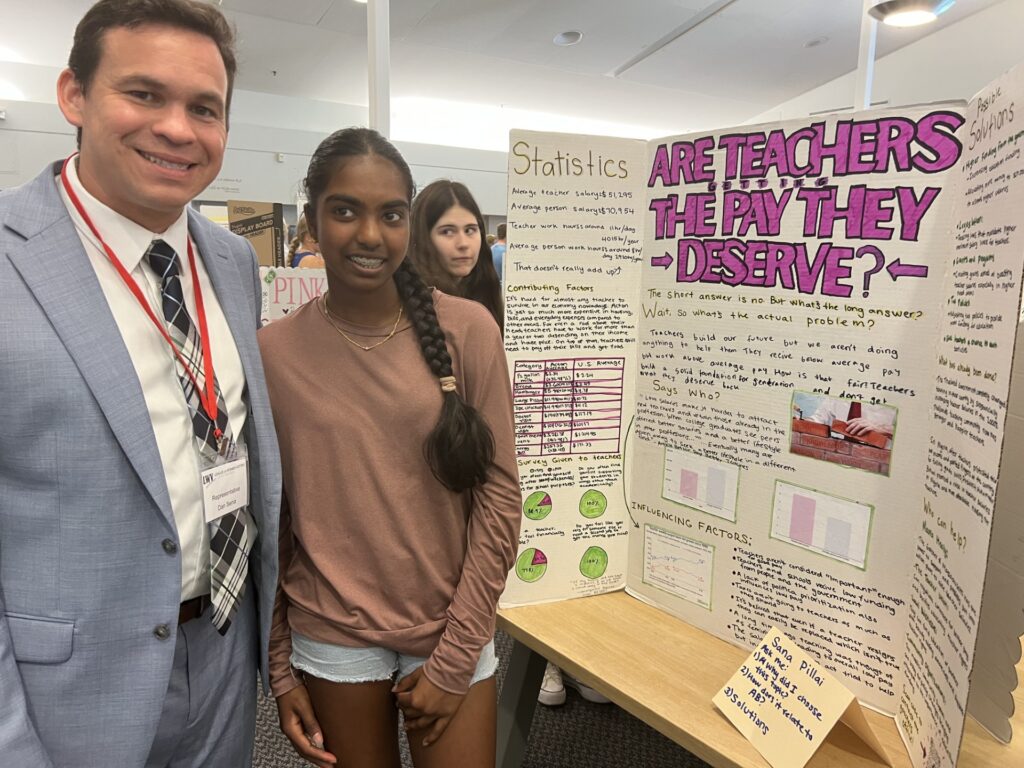 A man in a suit stands next to a girl with braces and a long braid. They are in front of a poster that askes, "Are teachers getting the pay they deserve?"