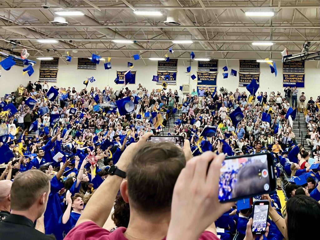 A chaotic picture with people taking photos in the foreground, a swilrl of blue gowns in the middle ground, and many mortarboards at the top of the photo.