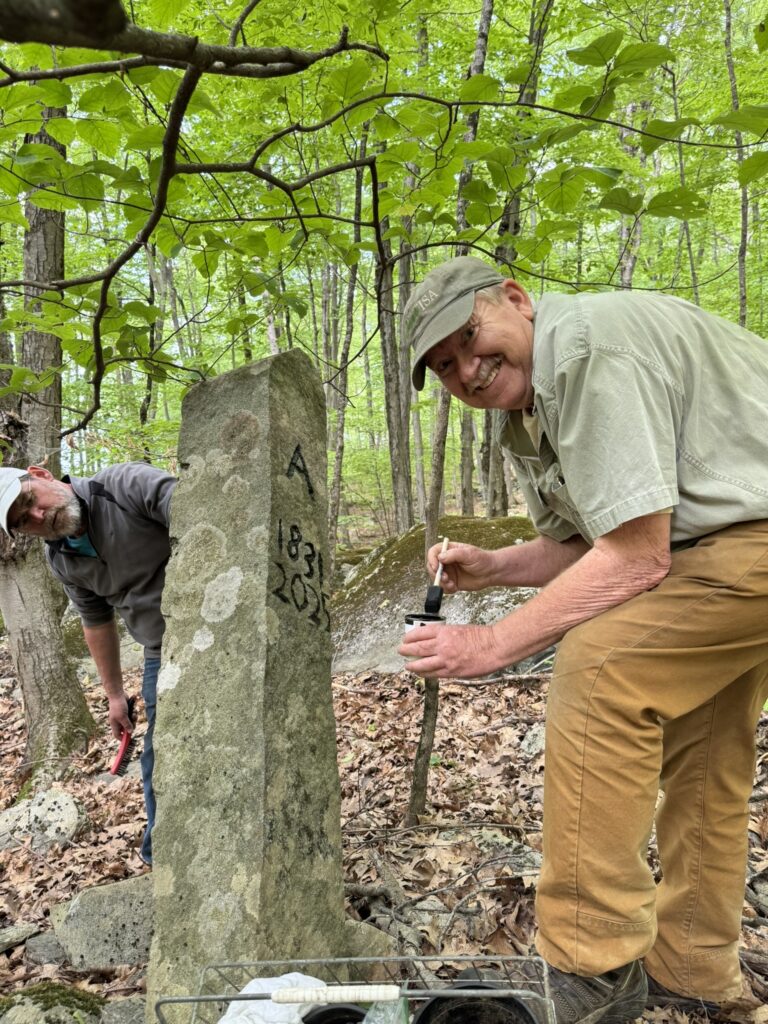 A man wearing a green cap paints the letter A (for Acton) and the numbers 1831 - 2025 on a stone pillar. On the other side of the pillar another man is marking the Littleton side.