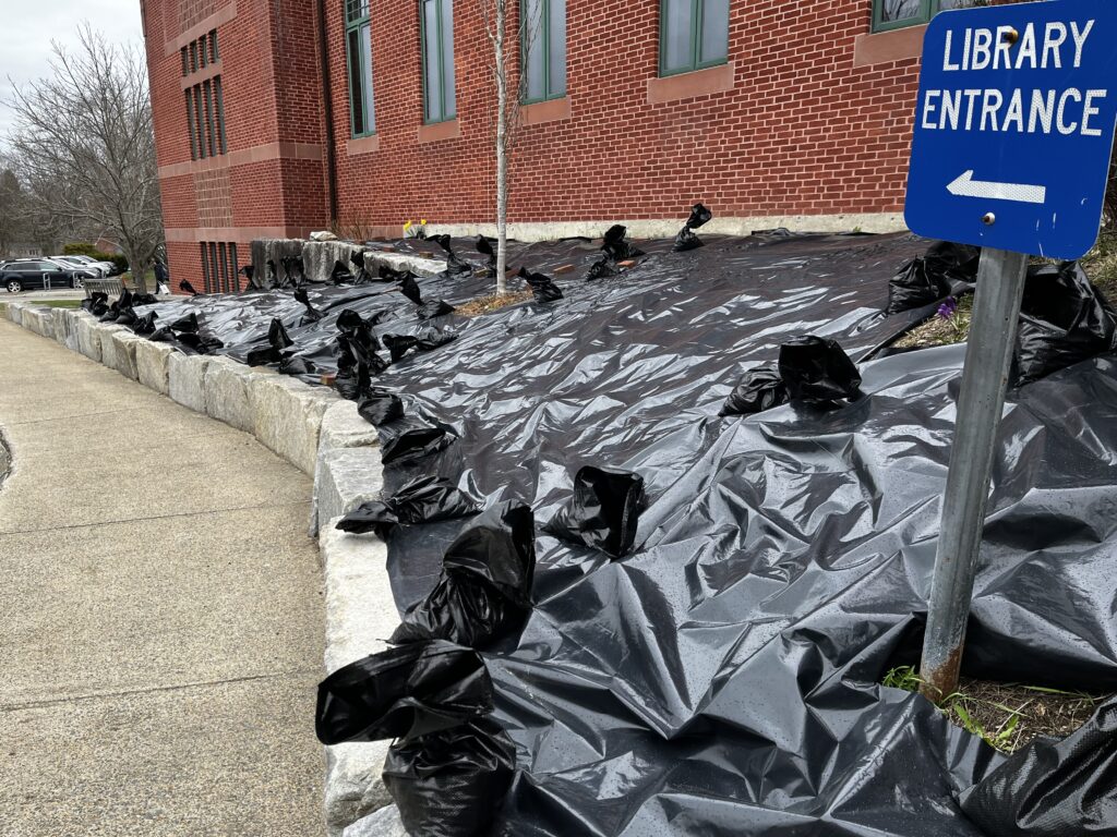 Undereneath a brick wall, an area covered by a black plastic tarp. The Library Entrance sign pokes out of a plastic sheet.