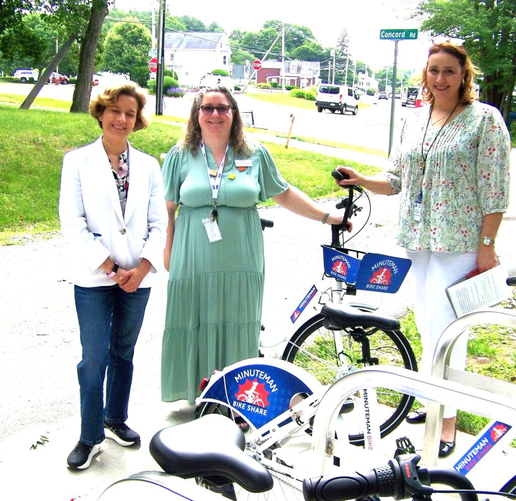 Three women stand out in the sunligh with Minuteman Bike Share bikes.