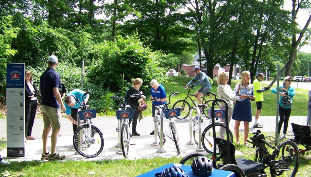 One person riding a bike, another person adjusting a bike, and more people standing around the bikes and chatting.