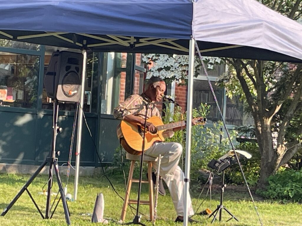 A Black man playing a guitar under a canopy on a lawn. The sun is low enough that you can clearly see his face.