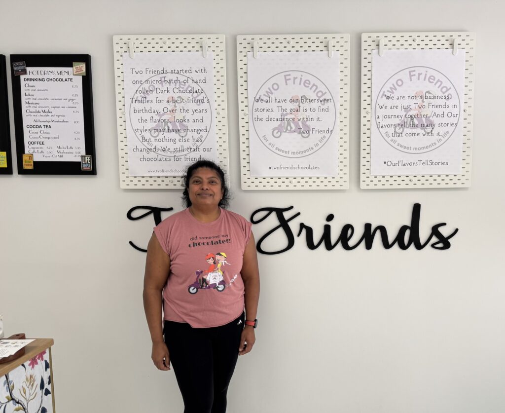A smiling woman wearing a "Did someone say chocolate" t-shirt stamds in front of a series of signs inside the Two Friends store.