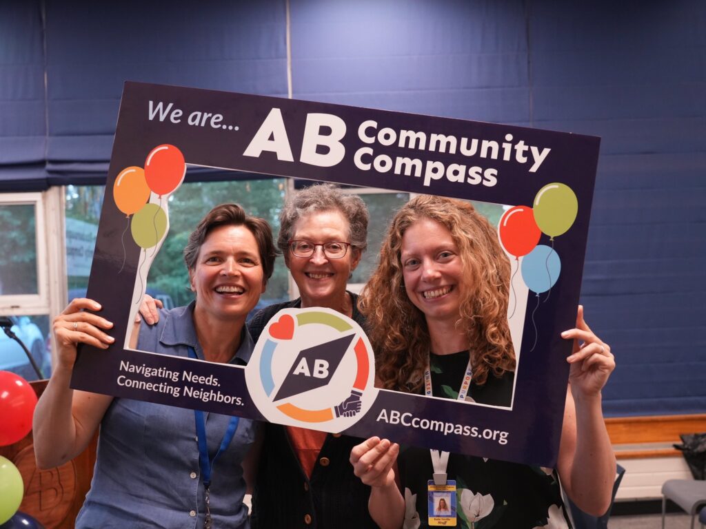 Three women stand behing a frame that says "We are.... AB Community Compass."