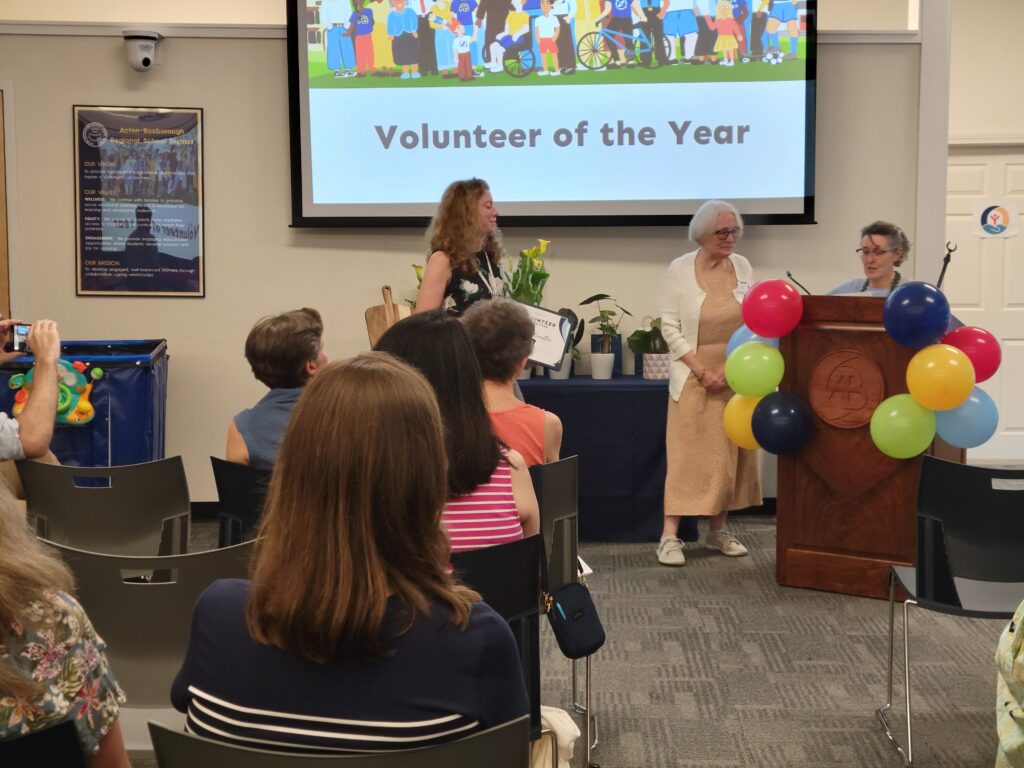 A woman in a yellow dress stands under a screen that proclaims "Volunteer of the Year." On the left, a woman in a black dress holds flowers, and on the right, a woman speaks behind a balloon-covered podium.