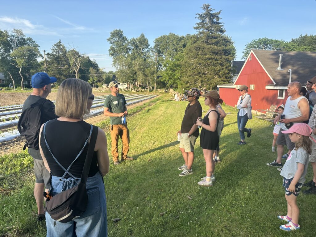 A man in a green BAG polo shirt speaks while several other people listen. They are out in a field with a red barn and black plastic-covered rows in the background.