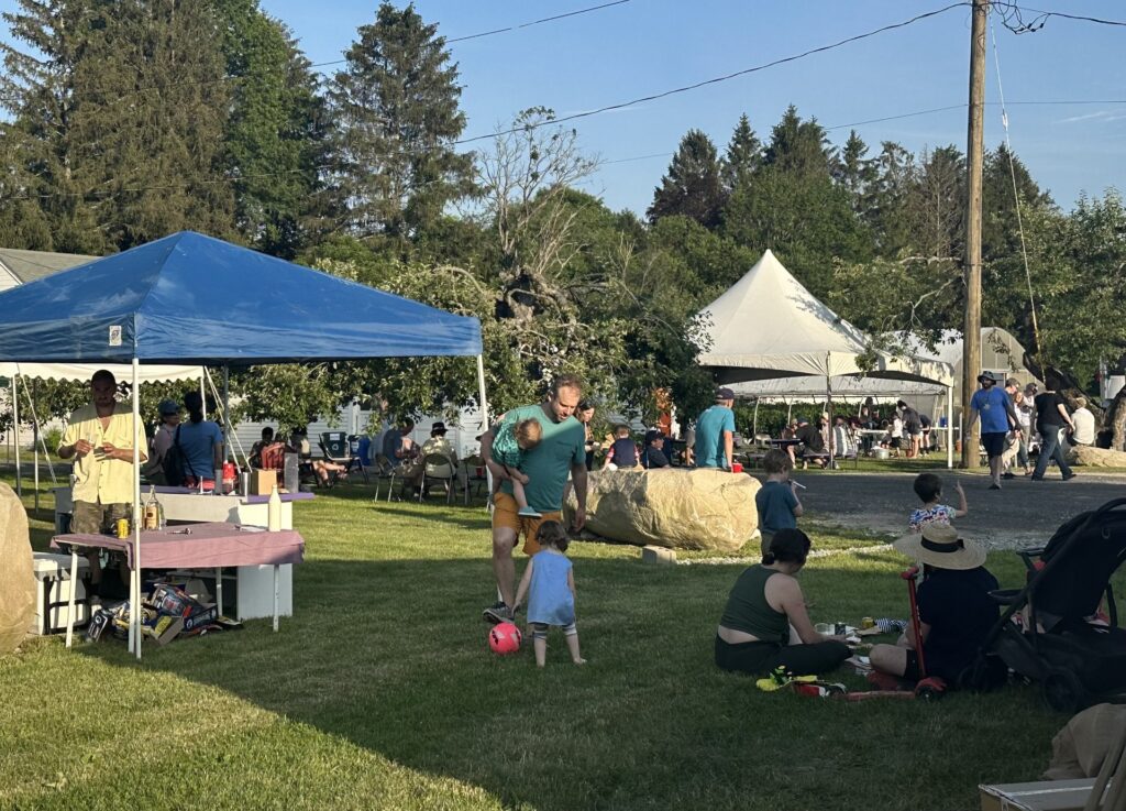 Tents on a lawn on a sunny evening. In the front, a small child plays with a ball, while the dad holds an even small child.