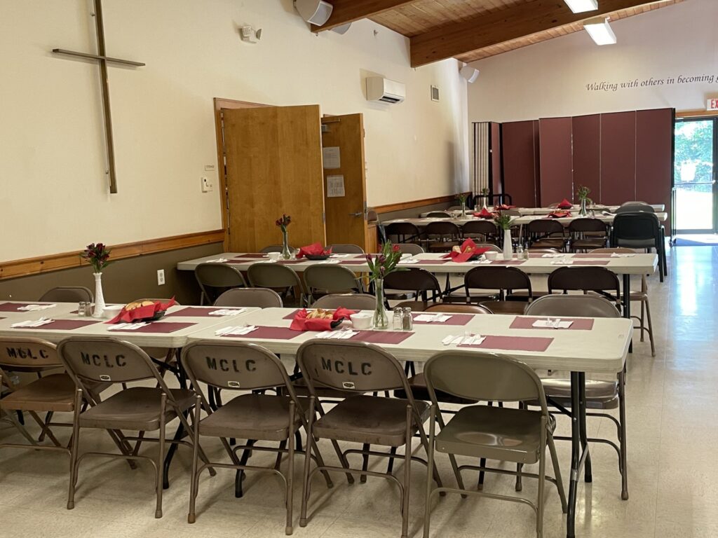A hall with tables set with placemats, plasticware, and baskets of bread. There are vases of flowers on each table.