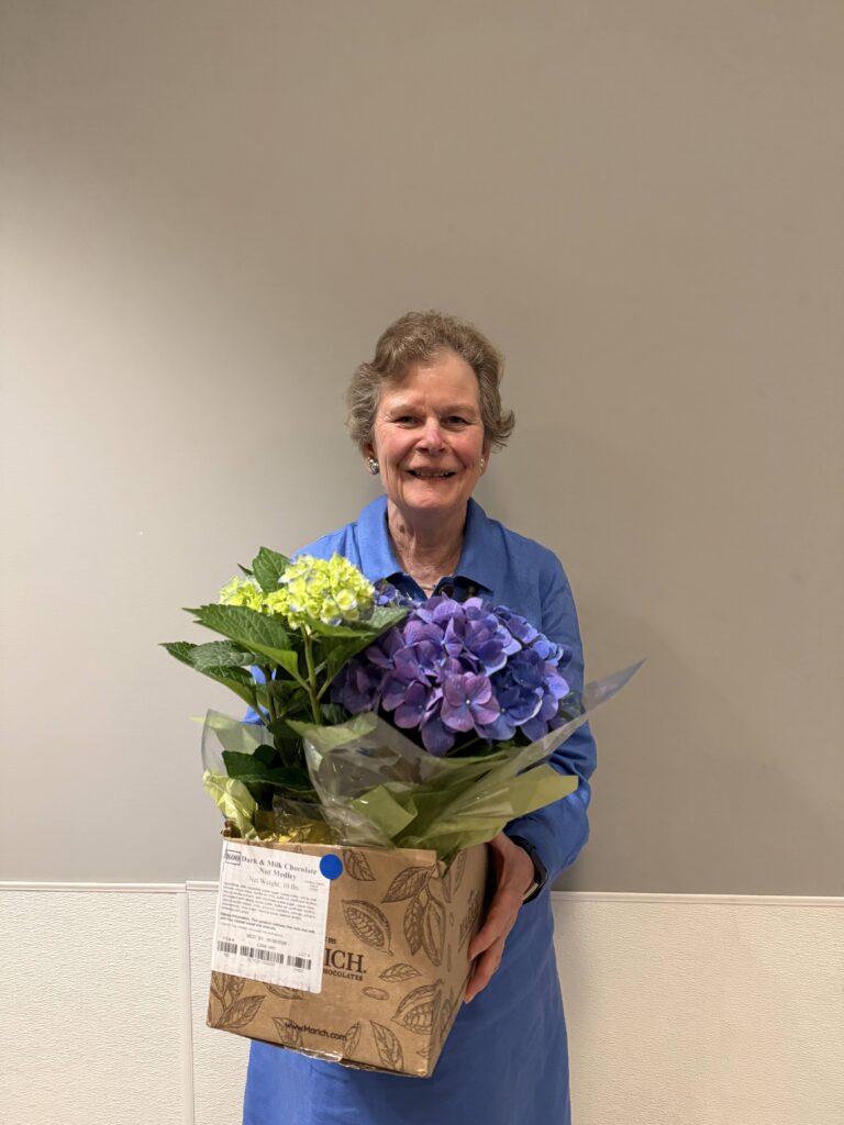 A smiling woman in a blue dress holds a large box of flowers.