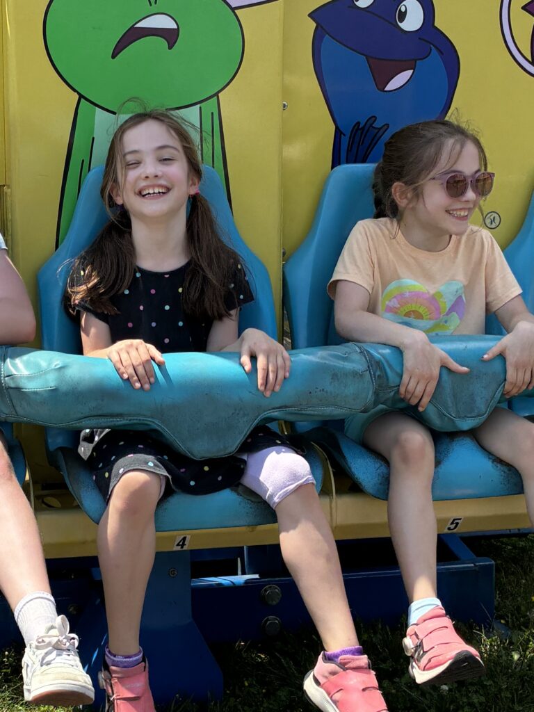 Twin girls smile broadly while locked into a ride with cartoon characters at their back.
