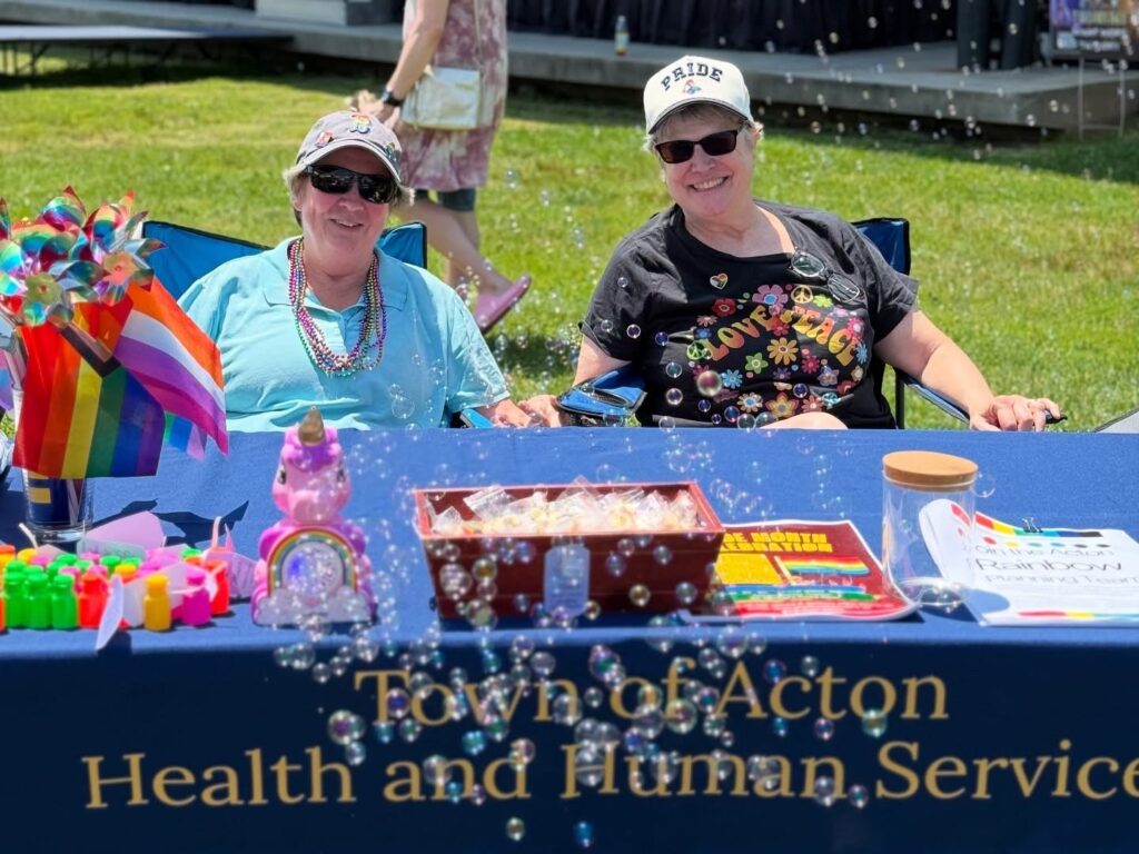 Two people sit behind the Acton Health and Human Services table, which has plenty of great swag. A bubble machine makes the picture a little bit fuzzy.