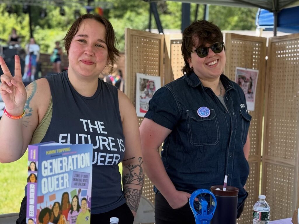 Two people under a tent. The person on the left is showing off their new book "Generation Queer."