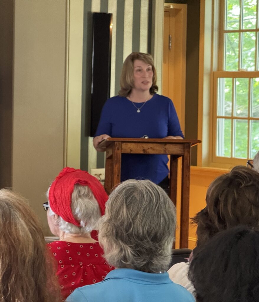 A blonde woman with a blue shirt stands at a desk speaking to an audience.