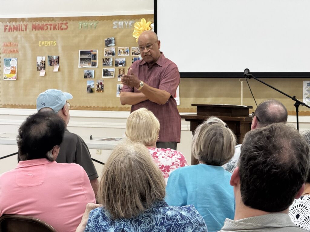 An older Black man stands in front of an audience. He is gesturing like he's answering a question.