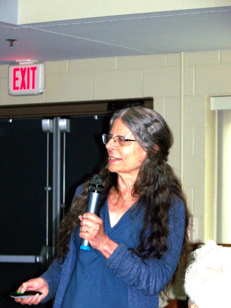 A woman with long hair, glasses, and a blue dress speaks into a mic.