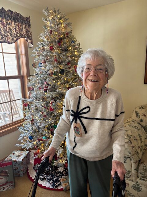 A smiling older woman stands in front of a Christmas tree. She's wearing a sweater that looks like a package.