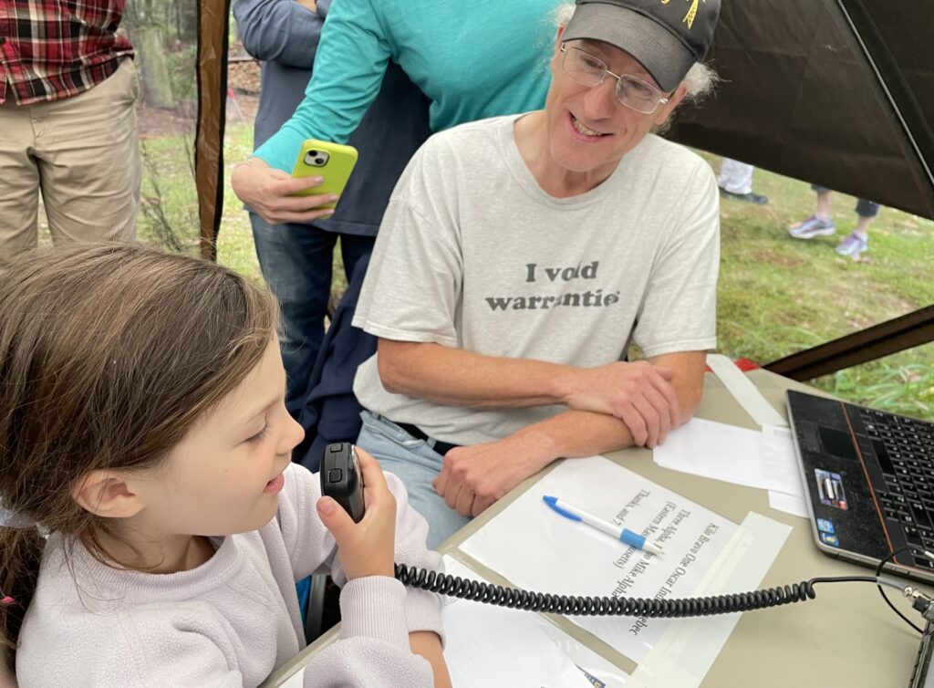 A young girl speaks into a radio mic. She's sitting with a man wearing a baseball cap and a t-shirt that says, " I void warranties."