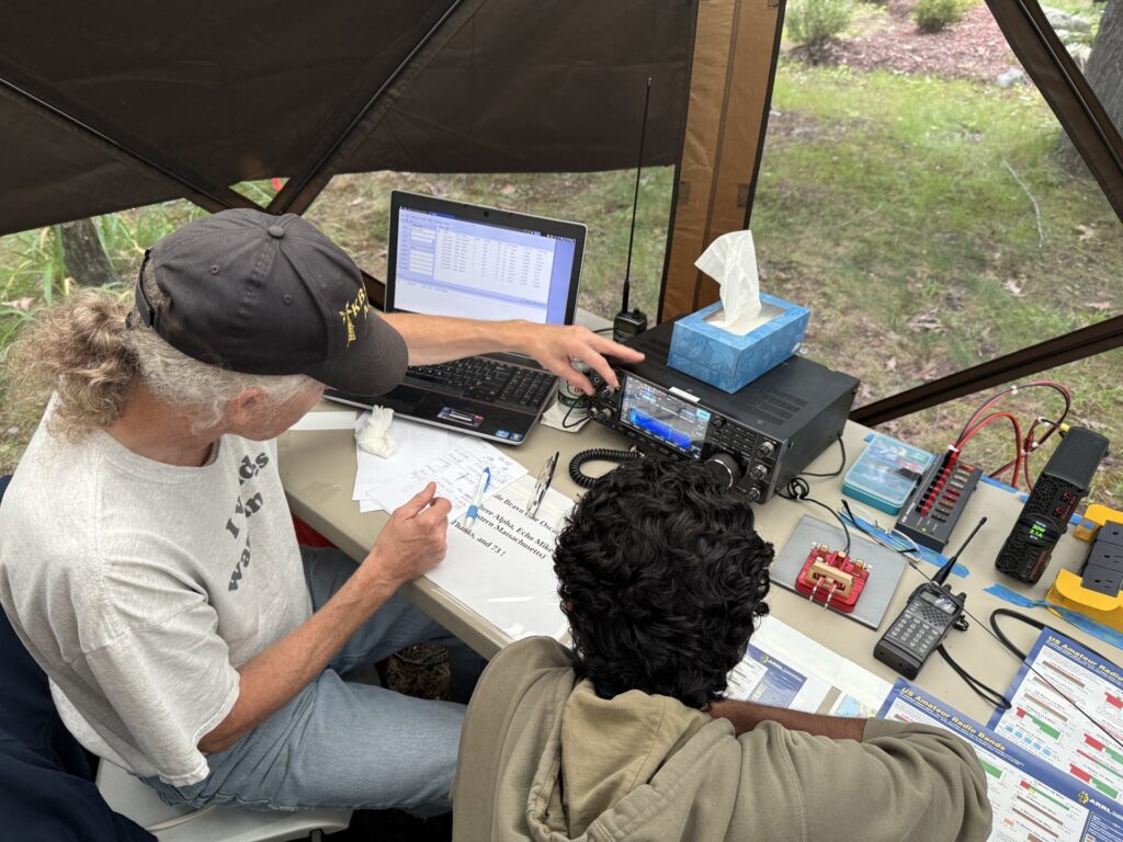 An older man and a younger man sitting at a desk. The older man is showing the younger one some radio equipment.
