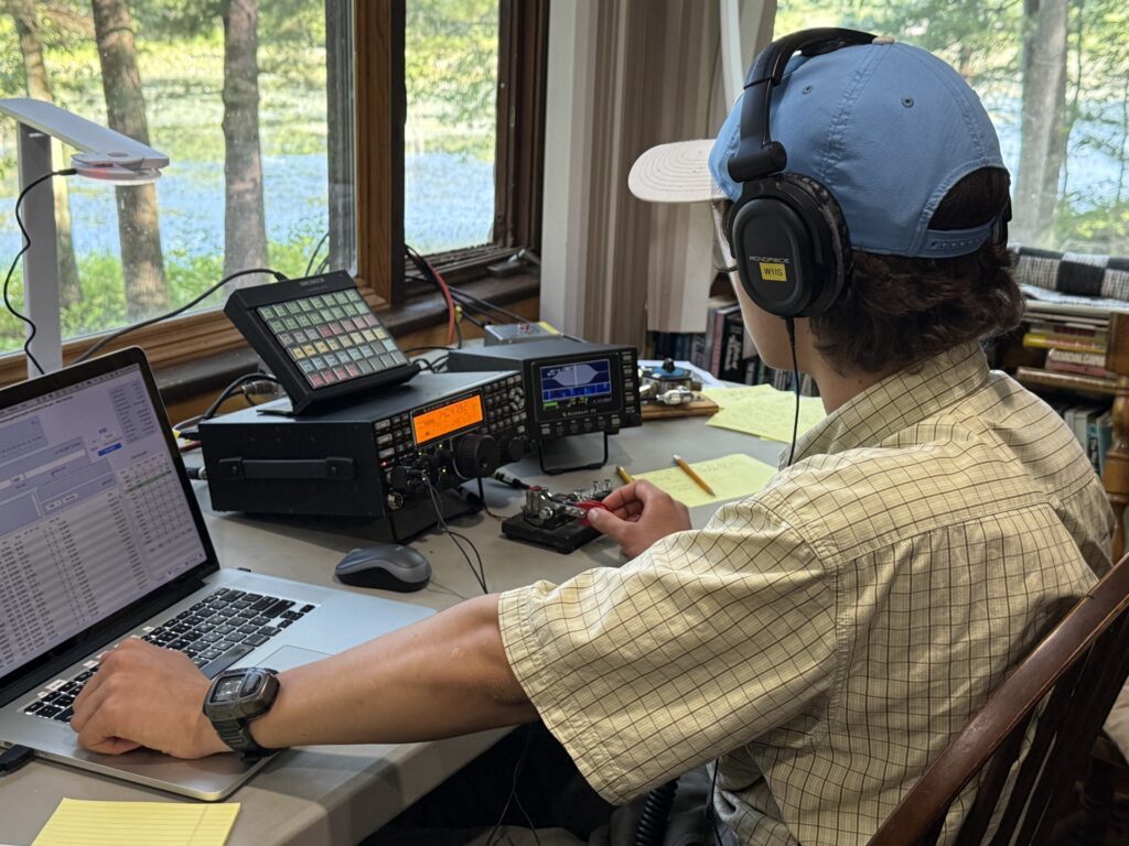 A man sits at a desk. His left hand is on a computer, his right hand is on a Morse code generator.
