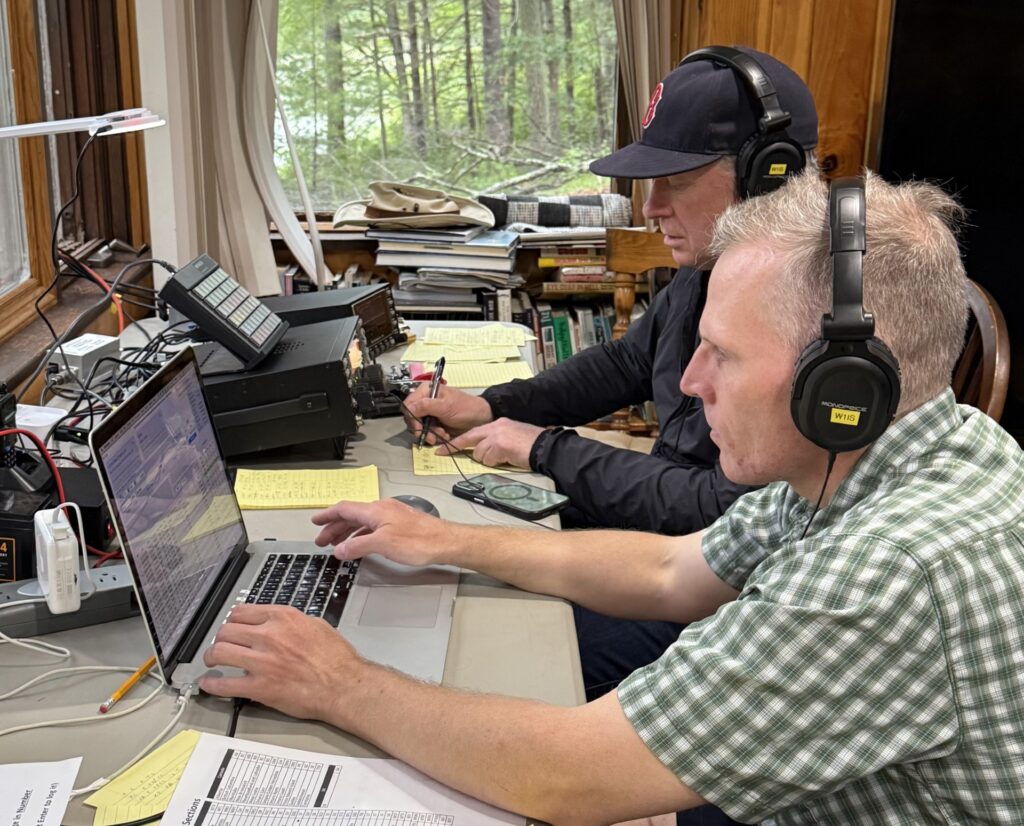 Two men sit at a desk. One is in front of a computer, the other has a paper and pen (to interpret Morse code?).
