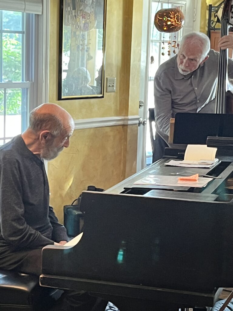 An older man, balding and with a grey beard, sits at a piano. Behind him, a bass player leans over to talk to him.