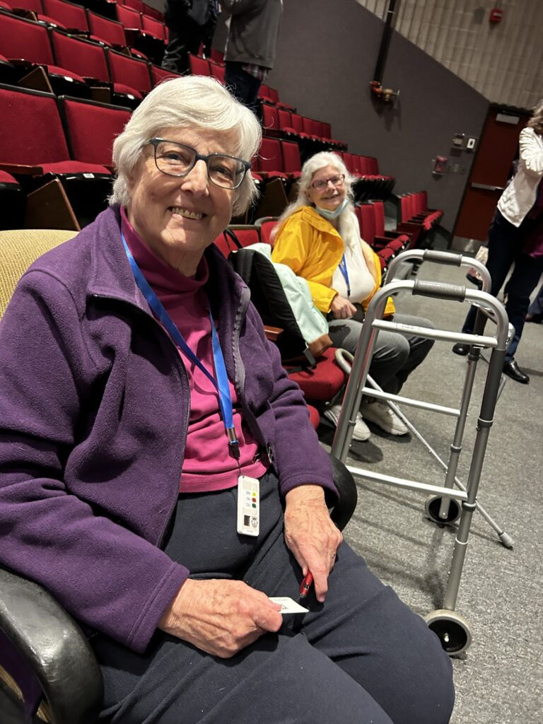 Two women sitting in an auditorium. The woman in the front has short white hair, glasses, and a wise smile. The woman in the back, with long white hair is also smiling.