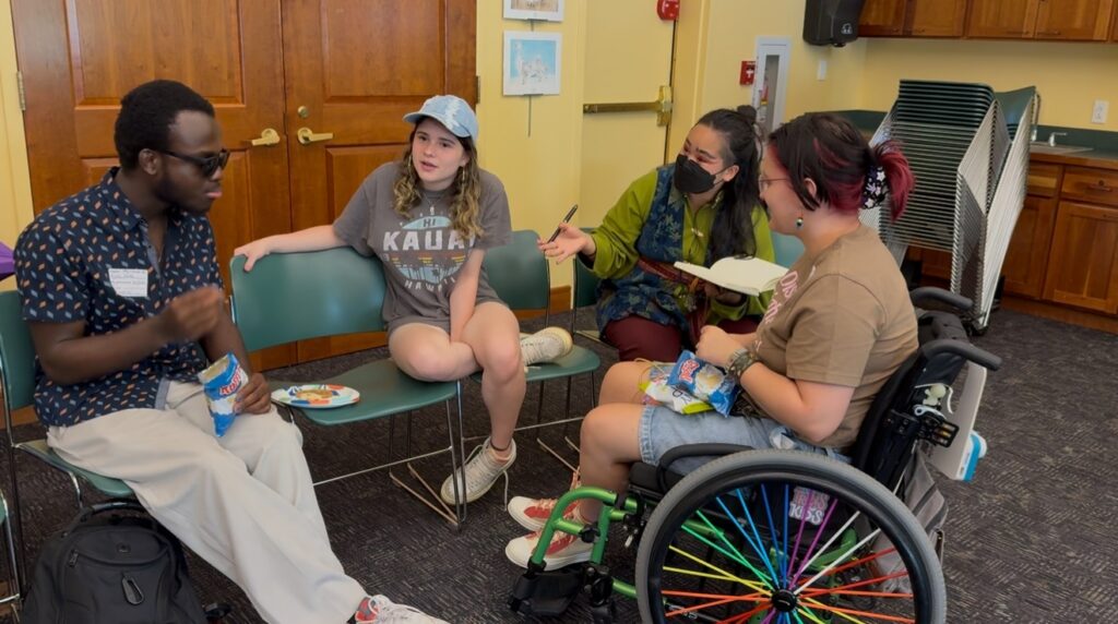 Three people sit in chairs in a circle. A fourth person is part of the circle in a wheelchair with rainbow spokes.