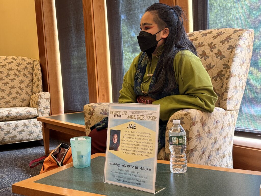 A person with exotic eye makeup and a black mask sits in a chair.
