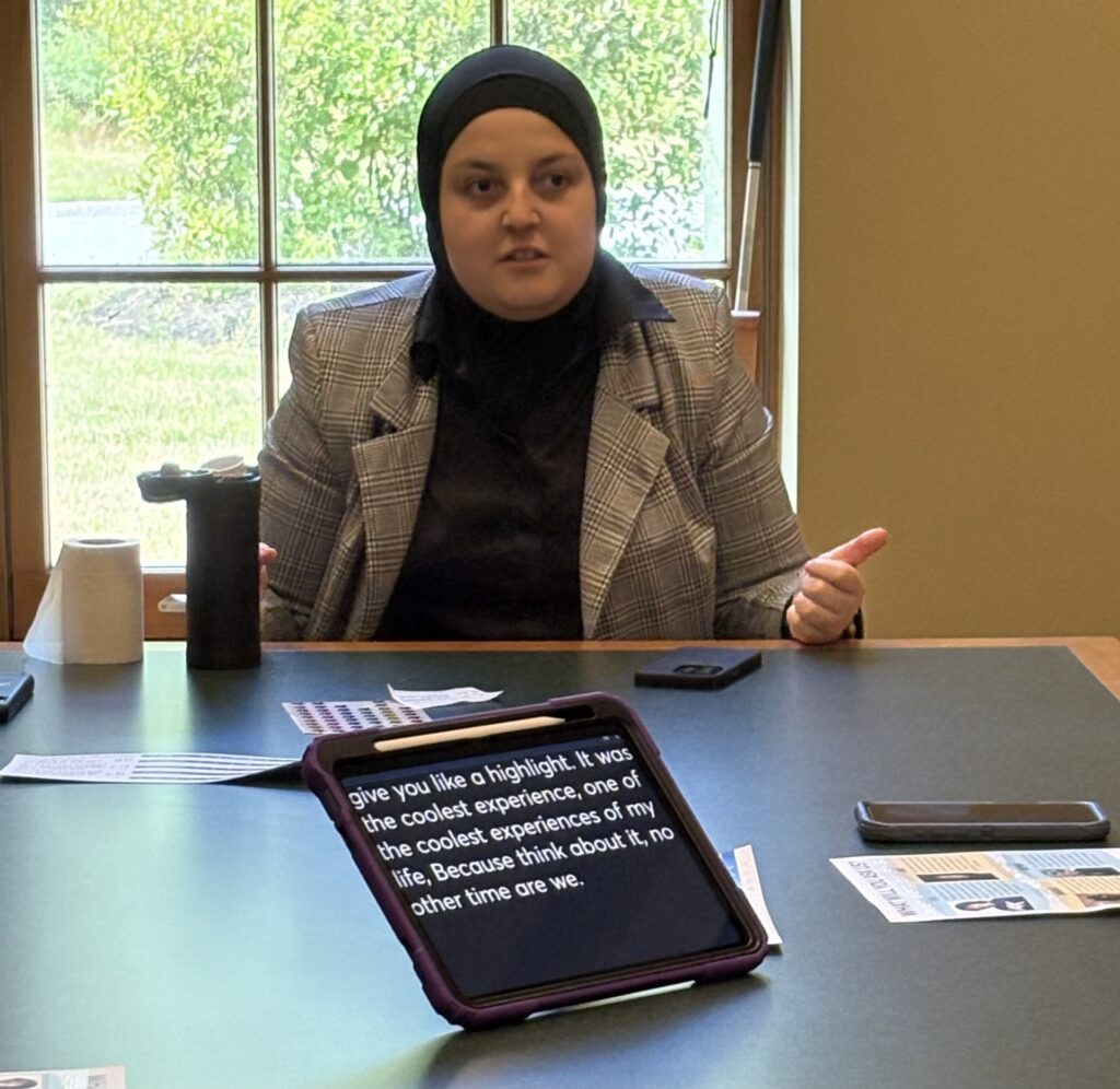 A woman sits at a desk. On the desk, an iPad-like device transcribes the words that she is saying.
