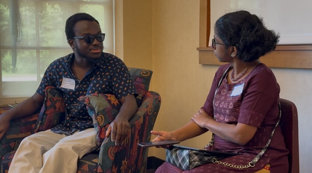A man wearing sunglasses speaks with a young woman. She has a phone in her hand and an iPad on her lap (for audio transcription).
