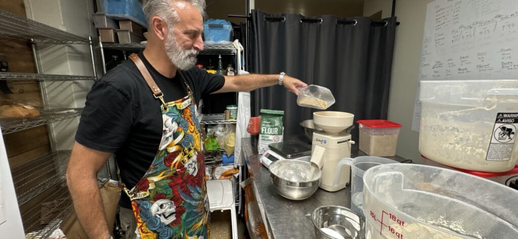 A tall bearded man wearing an apron pours grain into a small grain mill. In the background is a cooling rack, an 18 quart dough bucket, and other baking accoutrements.