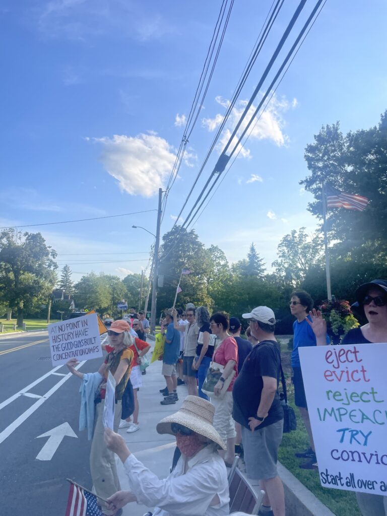 People lining th stret on a hot summer day. Some are carrying posters and Amercan flags. One poster says "At no point in history have the good guys built concentration camps."