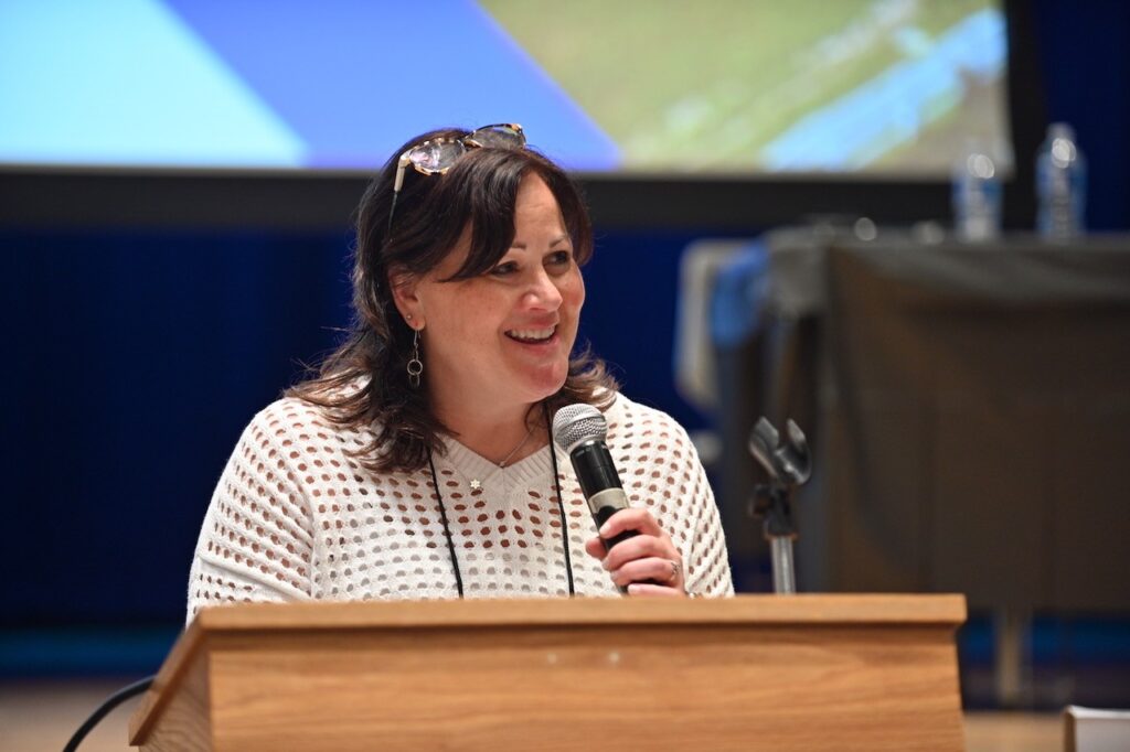 A woman with sholder length black hair and glasses propped on top of her head speaks into a microphone.