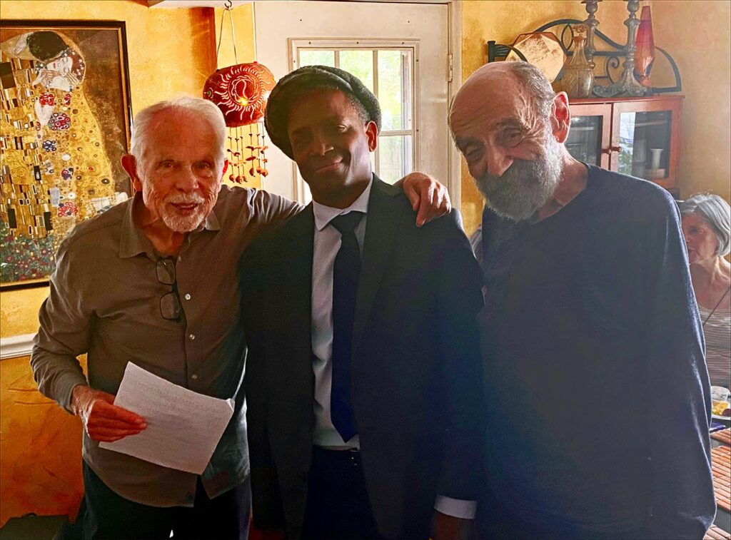 Three men stand in front of a wall with a Klimt print in the background. Two of the men are older, the drummer (in the middle) is much younger.