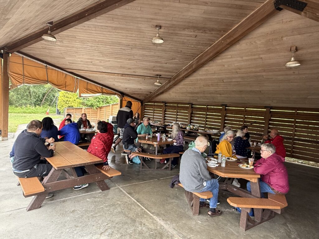 People at picnic tables underneath an open-sided wooden structure. It looks rainy outside.