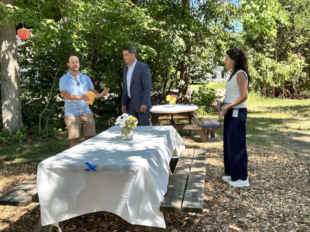 Three people stand talking behind a picnic table. One of the men is speaking.
