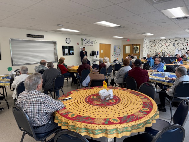 People sit around round tables with nice tablecloths. They are facing a man standing at the front of the room.