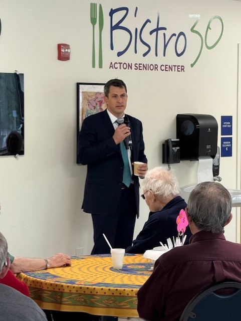 A man in a suit and tie stands in the Acton Senior Center. One hand holds a mic, the other, a cup of coffee.